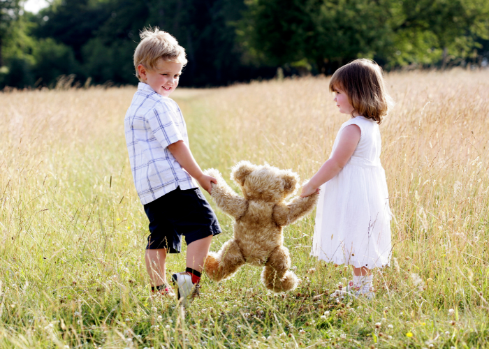 Little boy and little girl holding teddy bear walking through field.